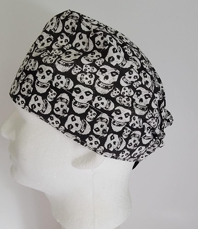 May include: A black and white skull print scrub cap. The cap is made of fabric and is designed to cover the head. The skull pattern is repeated across the entire surface of the cap. The cap is shown on a white mannequin head.