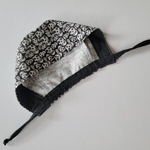 May include: A black and white skull print surgical cap. The cap features a repeating pattern of white skulls on a black background. The interior lining is a lighter gray with a subtle pattern. It has black ties for securing.
