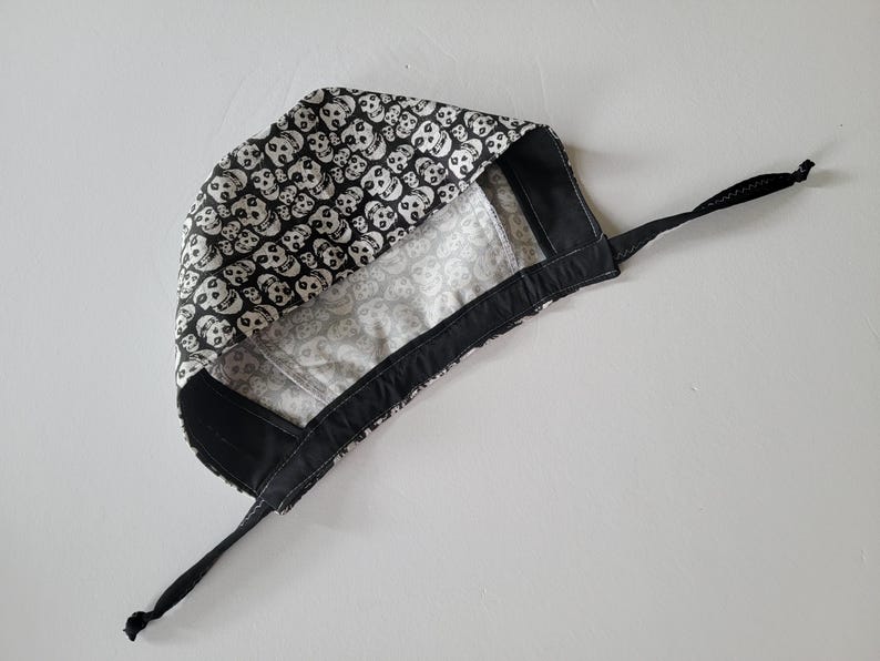 May include: A black and white skull print surgical cap. The cap features a repeating pattern of white skulls on a black background. The interior lining is a lighter gray with a subtle pattern. It has black ties for securing.