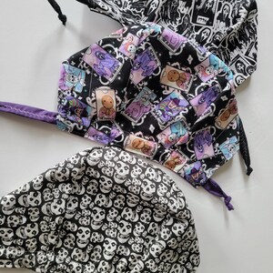 May include: Three Halloween-themed scrub caps. One cap features a black and white pattern of ghosts and the text "1996". Another cap has a black background with colorful illustrations of spooky characters. The third cap is covered in a pattern of white skulls on a black background.