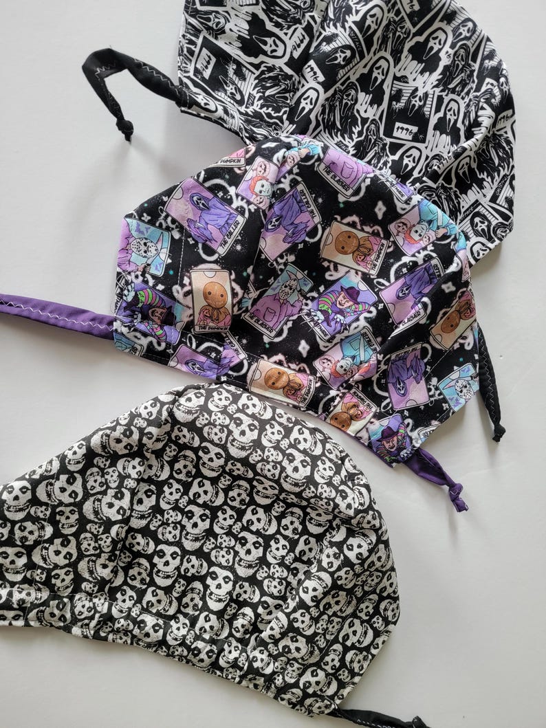 May include: Three Halloween-themed scrub caps. One cap features a black and white pattern of ghosts and the text "1996". Another cap has a black background with colorful illustrations of spooky characters. The third cap is covered in a pattern of white skulls on a black background.
