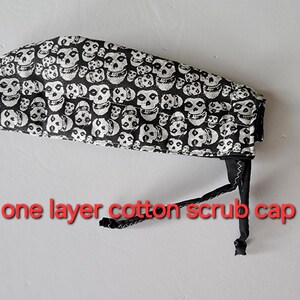 May include: Black and white cotton scrub cap featuring a repeating skull pattern. The cap has a black tie closure and is labeled "one layer cotton scrub cap" in red text. This medical-themed accessory is designed for healthcare professionals.