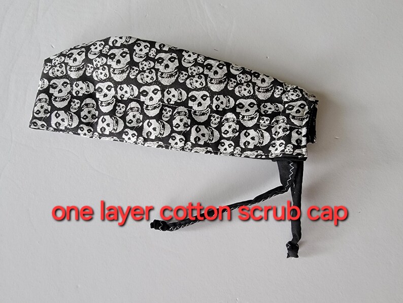 May include: Black and white cotton scrub cap featuring a repeating skull pattern. The cap has a black tie closure and is labeled "one layer cotton scrub cap" in red text. This medical-themed accessory is designed for healthcare professionals.
