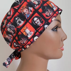 May include: A red and black surgical scrub hat with a pattern of horror movie characters. The hat has a tie at the back.