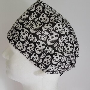 May include: A black and white skull-patterned surgical cap. The cap is made of fabric and features a repeating pattern of white skulls on a black background. The cap is designed to cover the head and hair.