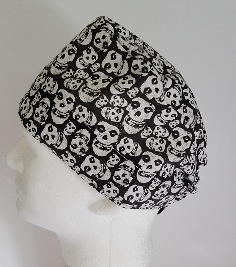May include: A black and white skull-patterned surgical cap. The cap is made of fabric and features a repeating pattern of white skulls on a black background. The cap is designed to cover the head and hair.