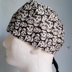May include: A black and white skull print cap, likely a bandana or head covering. The cap is patterned with numerous white skulls on a black background. The cap is tied at the back with a black tie.