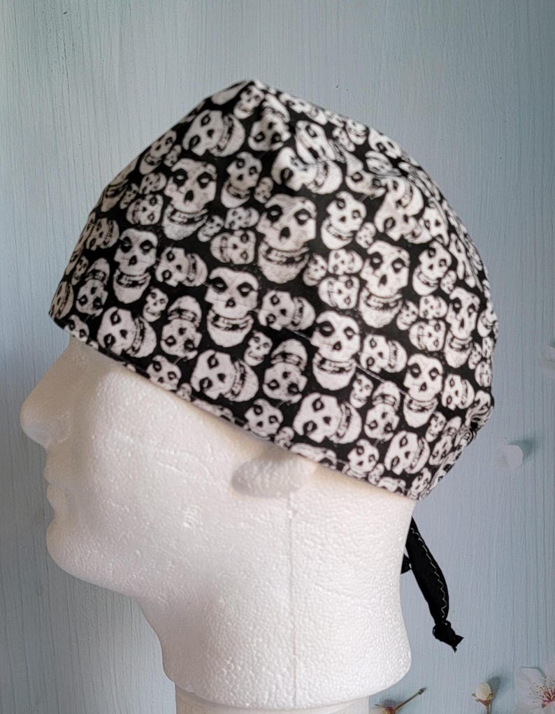 May include: A black and white skull print cap, likely a bandana or head covering. The cap is patterned with numerous white skulls on a black background. The cap is tied at the back with a black tie.