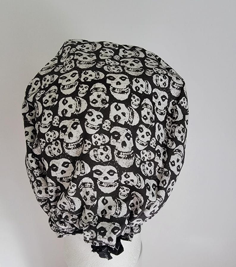 May include: A black and white skull print bouffant style cap. The cap is covered in a repeating pattern of white skulls on a black background. The cap is gathered at the base and appears to be made of fabric.