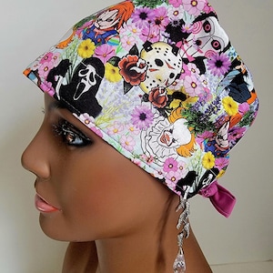 May include: A pink and white patterned surgical scrub cap featuring floral designs and horror movie characters. The cap includes images of Chucky, Jason Voorhees, and Pennywise. The cap has a pink tie in the back.