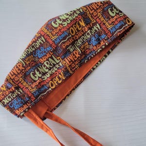 May include: A brown scrub hat with orange ties. The hat is printed with colorful medical terms, including "General Surgery", "Open", "Liver", "Resection", "Laparo", "Cholecystectomy", "Thyroid", "Hemorrhoids", "Stoma", and "Hernia".