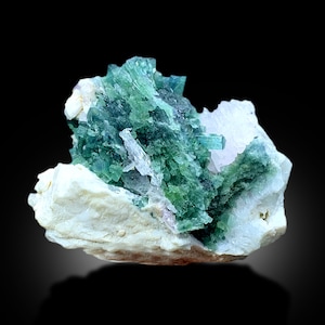 May include: A cluster of green and blue crystals on a white matrix. The crystals are sharp and pointed, and they are arranged in a random pattern. The matrix is rough and textured.