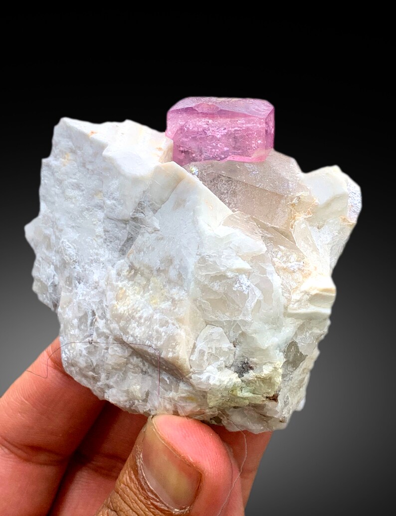 May include: A pink tourmaline crystal on a white matrix. The crystal is a cube shape with a smooth, shiny surface. The matrix is rough and textured.