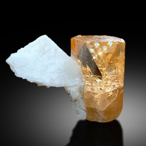 May include: A light orange topaz crystal with a clear, faceted, prism-shaped form. The crystal is resting on a white, opaque, mineral matrix.
