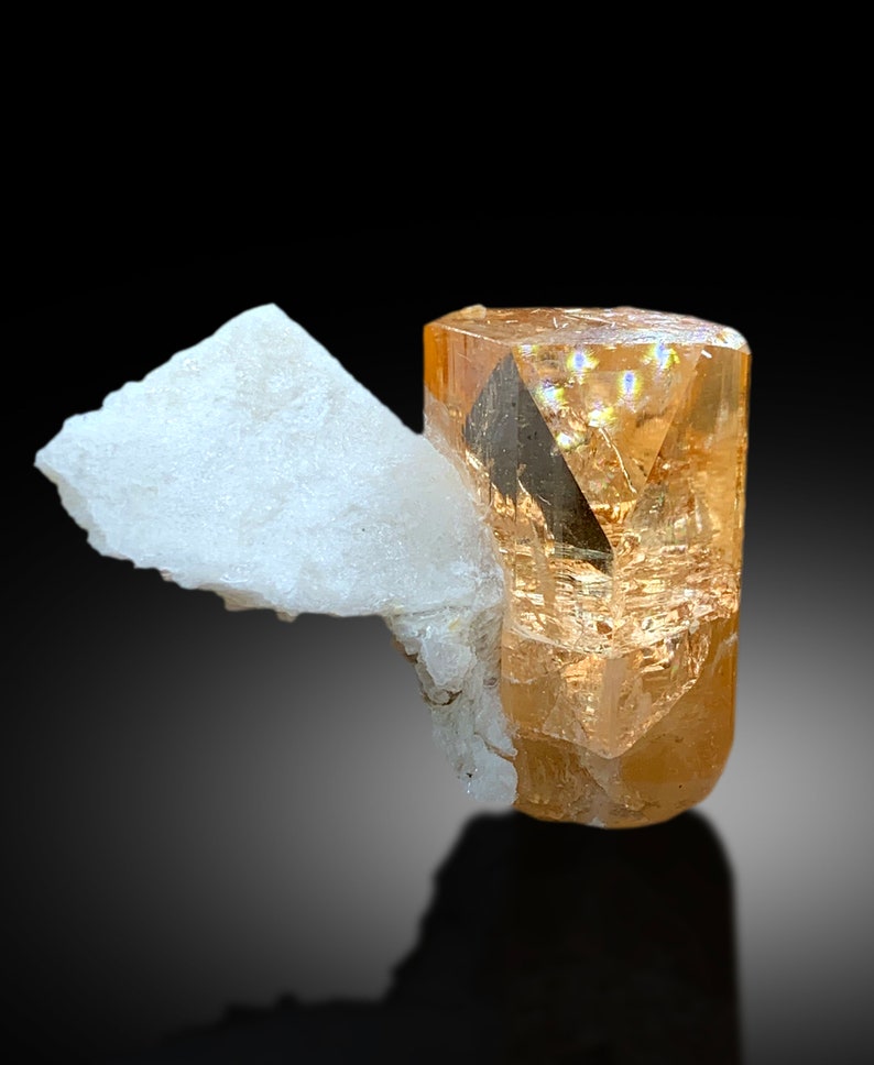 May include: A light orange topaz crystal with a clear, faceted, prism-shaped form. The crystal is resting on a white, opaque, mineral matrix.