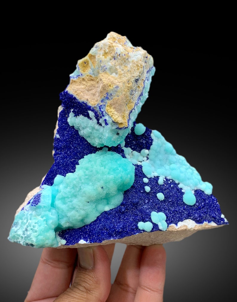 May include: A cluster of blue azurite and green chrysocolla crystals on a matrix of light brown rock. The crystals are arranged in a random pattern, with some of the azurite crystals forming small, rounded shapes.