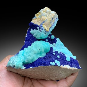 May include: A blue and green mineral specimen with a rough, natural texture. The specimen has a layered appearance, with a blue layer on top of a green layer. The specimen is sitting on a brown surface.