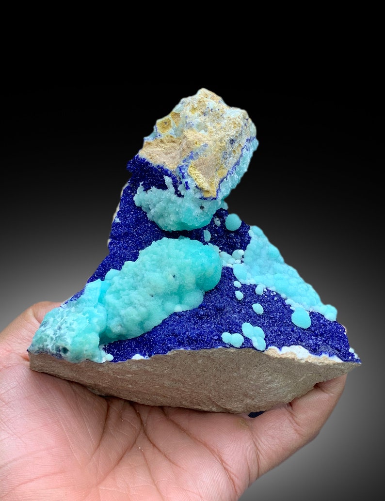 May include: A blue and green mineral specimen with a rough, natural texture. The specimen has a layered appearance, with a blue layer on top of a green layer. The specimen is sitting on a brown surface.
