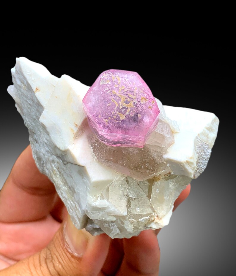 May include: A pink tourmaline crystal with a hexagonal shape, perched atop a white matrix of quartz crystals.