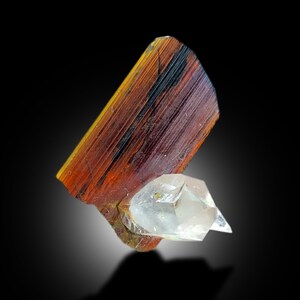 Op de afbeelding: Een close-up van een rood en oranje kristal met een helder kwartskristal eraan bevestigd. De kristallen bevinden zich op een zwarte achtergrond.