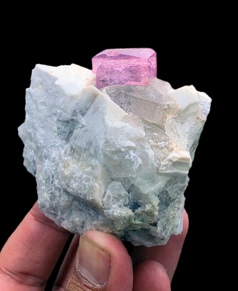 May include: A pink crystal, likely a tourmaline, sits atop a white and gray rock matrix. The crystal is well-formed and has a sharp, geometric shape.
