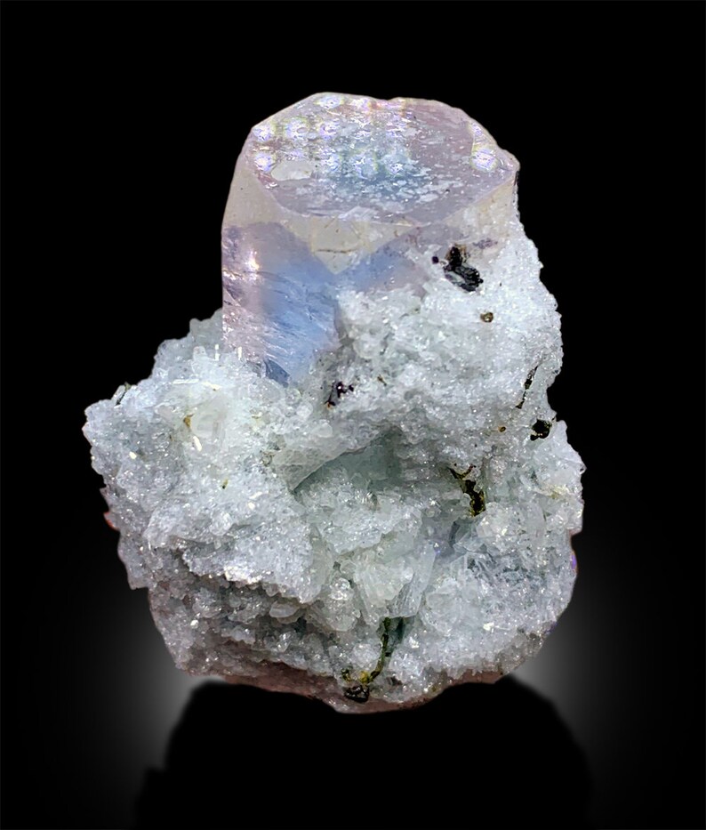 Puede incluir: Un cristal azul claro iridiscente de forma hexagonal, rodeado de una matriz de cristales blancos m&aacute;s peque&ntilde;os.