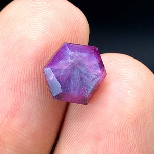 Puede incluir: Una piedra preciosa púrpura con forma hexagonal y corte facetado. La piedra preciosa tiene un tono púrpura claro con vetas púrpuras más oscuras.