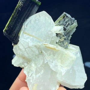 May include: A cluster of clear quartz crystals with two green tourmaline crystals on top. The quartz crystals are translucent and have a glassy luster. The tourmaline crystals are dark green and have a striated surface.