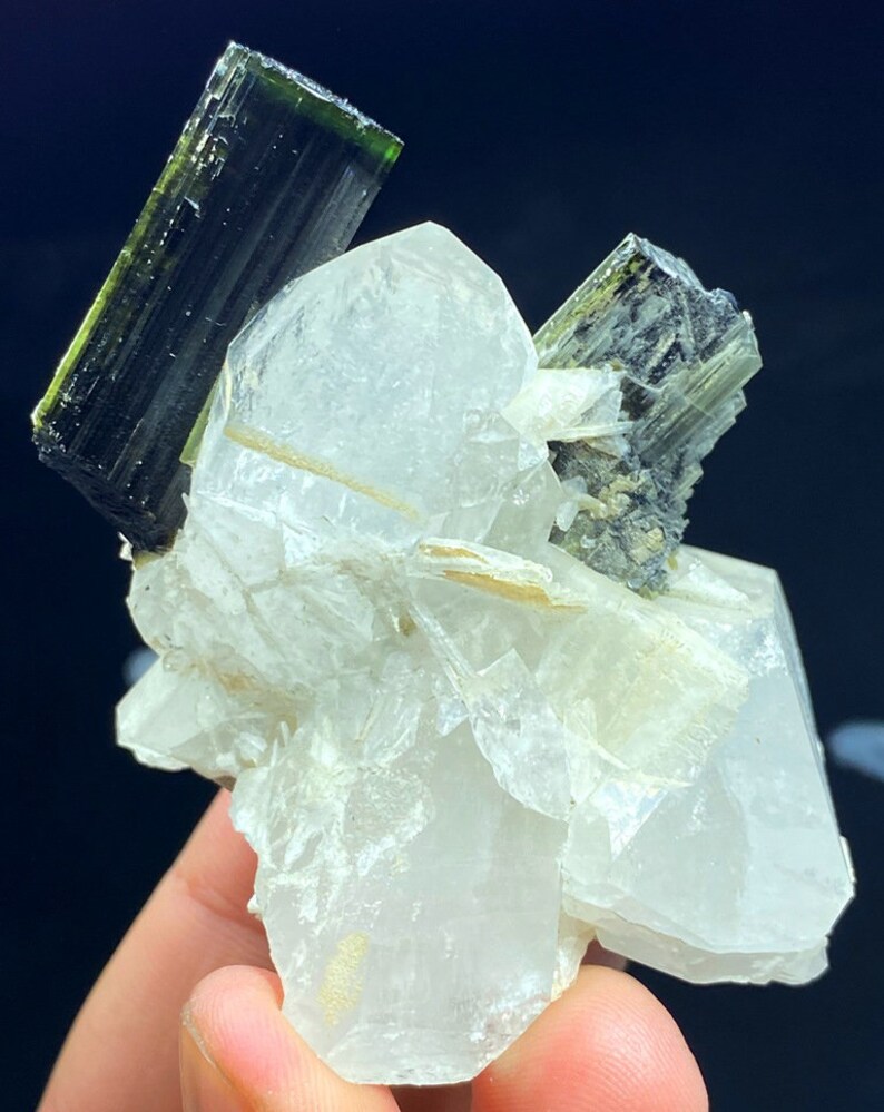May include: A cluster of clear quartz crystals with two green tourmaline crystals on top. The quartz crystals are translucent and have a glassy luster. The tourmaline crystals are dark green and have a striated surface.