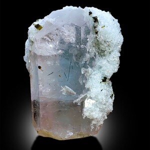 Puede incluir: Un cristal de aguamarina transparente, azul claro con un tono rosado, parcialmente cubierto de cristales blancos y verdes. El cristal mide aproximadamente 7,5 cm de alto.