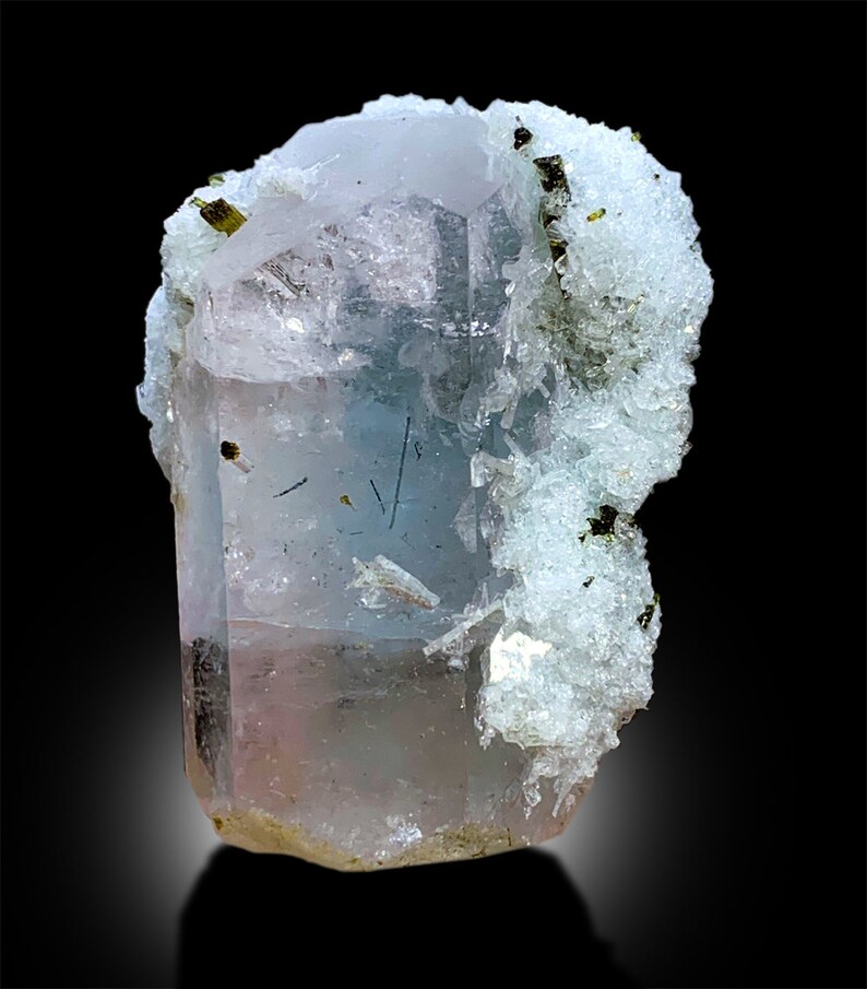 Puede incluir: Un cristal de aguamarina transparente, azul claro con un tono rosado, parcialmente cubierto de cristales blancos y verdes. El cristal mide aproximadamente 7,5 cm de alto.
