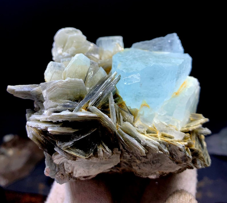 May include: A cluster of light blue aquamarine crystals on a bed of gray and white mica. The aquamarine crystals are translucent and have a glassy luster.