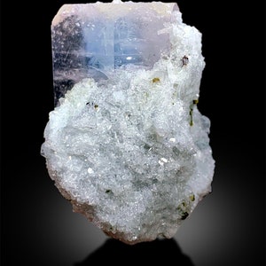 Puede incluir: Un gran cristal de aguamarina azul claro transparente con un tono rosado, incrustado en una matriz de cristales blancos y verdes.