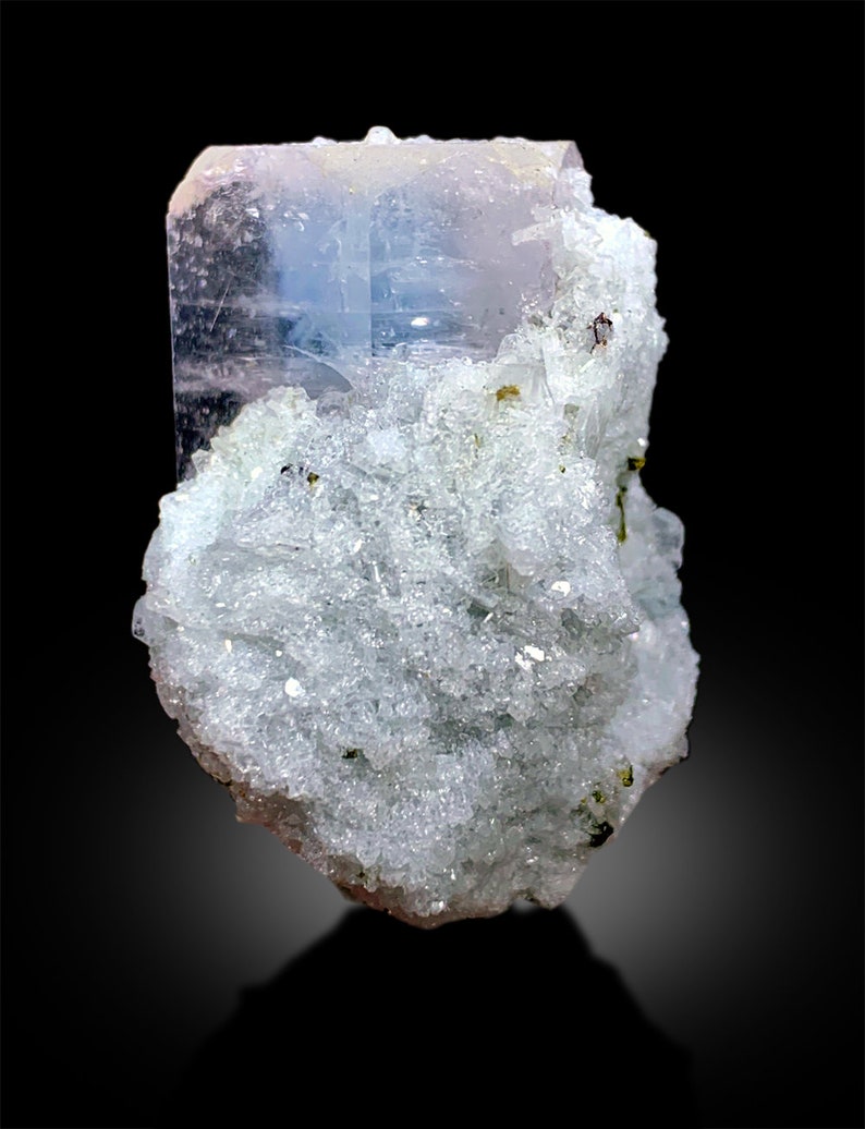 Puede incluir: Un gran cristal de aguamarina azul claro transparente con un tono rosado, incrustado en una matriz de cristales blancos y verdes.