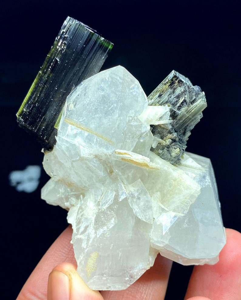 May include: A cluster of clear quartz crystals with two green and black tourmaline crystals. The quartz crystals are translucent and have a glassy luster. The tourmaline crystals are opaque and have a striated surface.
