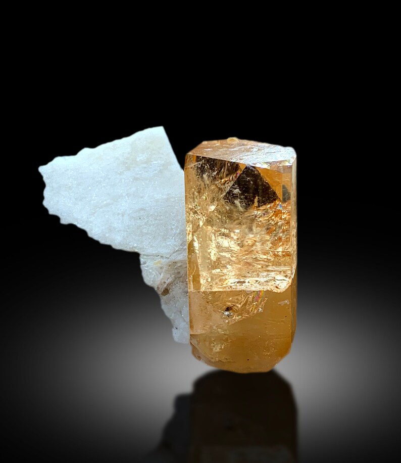 May include: A large, clear, orange-brown topaz crystal with a slightly cloudy appearance. The crystal is sitting on a piece of white rock.