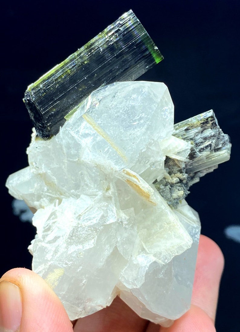 May include: A cluster of clear quartz crystals with two green and black tourmaline crystals on top. The quartz crystals are clear and have a glassy luster. The tourmaline crystals are green and black and have a striated surface.