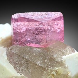 May include: A pink, translucent, rectangular crystal sits atop a cluster of white and clear quartz crystals.