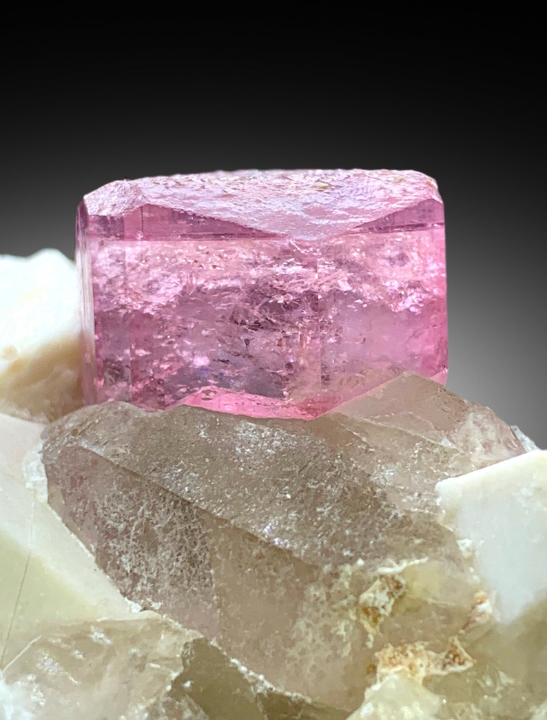 May include: A pink, translucent, rectangular crystal sits atop a cluster of white and clear quartz crystals.