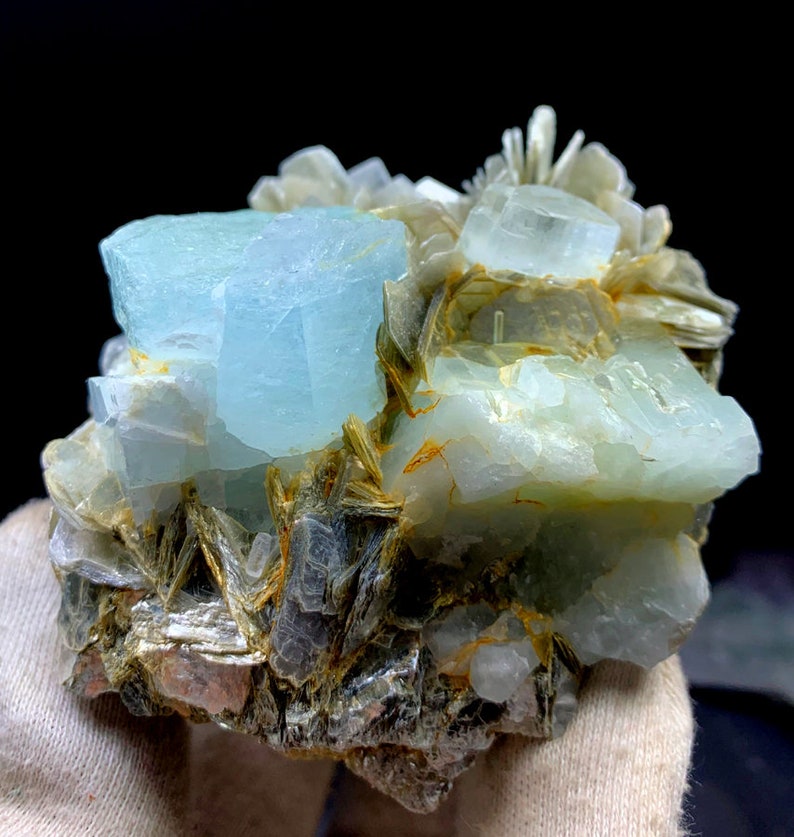 May include: A cluster of aquamarine crystals with a light blue color, embedded in a matrix of gray and brown mica. The crystals have a glassy luster and are translucent.