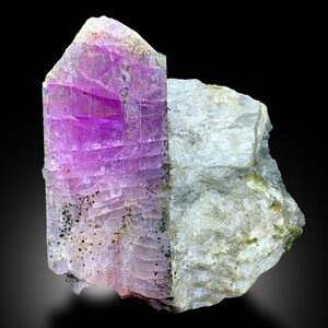 May include: A large, purple, and white crystal with a striated texture. The crystal is embedded in a gray, white, and green rock.