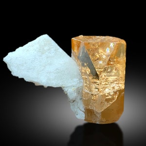May include: A clear, light orange topaze crystal with a hexagonal shape, sitting on a white, opaque mineral. The crystal is highly reflective and has a glassy luster.