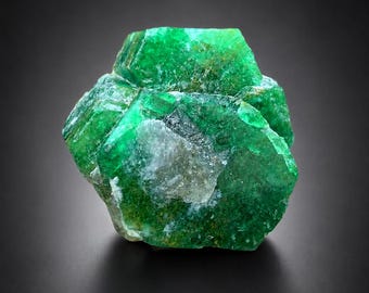 May include: A raw, hexagonal emerald crystal specimen featuring deep green coloration with translucent and opaque sections. The mineral displays natural crystalline facets, internal inclusions, and a rough, unpolished texture throughout its structure.