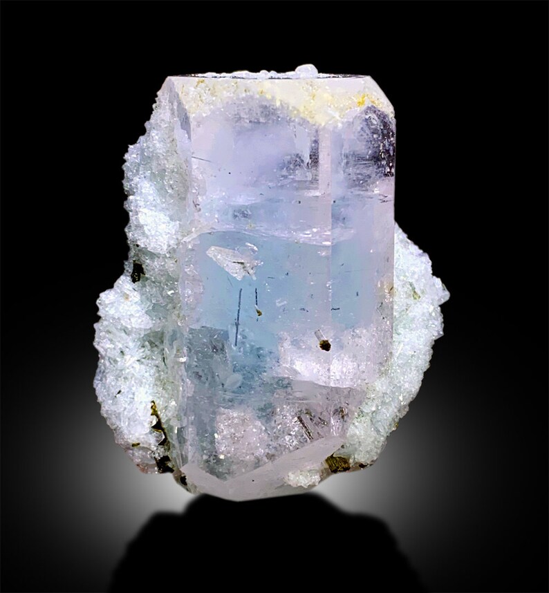 Puede incluir: Un cristal de aguamarina transparente, azul claro con un ligero tono rosado. El cristal est&aacute; en una matriz blanca rugosa.