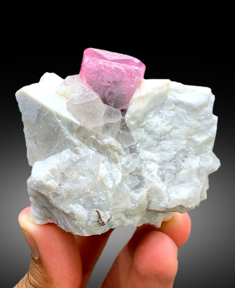 May include: A pink, translucent, faceted crystal sits atop a white, rough, rock matrix. The crystal is a vibrant pink color and has a sharp, geometric shape.