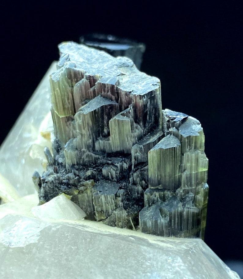 May include: A cluster of green and black tourmaline crystals on a clear quartz matrix. The crystals are sharp and well-defined, with a striated surface.