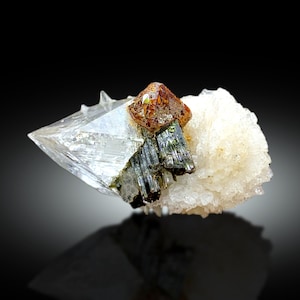 Puede incluir: Un cristal de cuarzo transparente con un cúmulo de minerales marrones y verdes en la parte superior. El cristal está sentado sobre un lecho de cristales blancos.