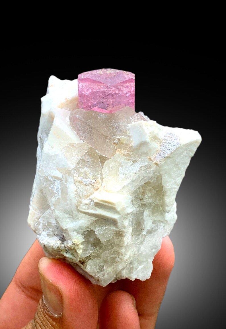 May include: A pink tourmaline crystal on a white matrix of quartz crystals. The tourmaline is a hexagonal prism with a smooth, glassy surface. The quartz crystals are clear and have a rough, textured surface.