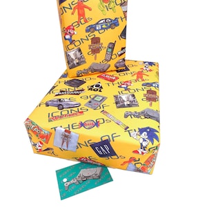 Puede incluir: Papel de regalo amarillo con un tema de los 90 con varios iconos de la d&eacute;cada, incluyendo videojuegos, dulces y un coche. El papel tiene el texto "Icons of the 90s" y "Gap" impreso en &eacute;l.
