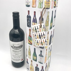 May include: A white wine bottle gift bag with a colorful pattern of various wine bottles. The bag has a brown twine bow and a small gift tag with a similar pattern. A bottle of red wine is next to the bag. The bottle label reads "Merlot Pays d'Oc 2013 Tanners Established 1945".